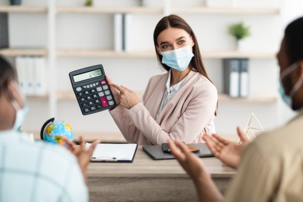Travel Agency Service. Female Agent Showing Calculator With Price Of Family Tour Consulting African American Tourists Couple In Modern Office, Wearing Face Mask. Selective Focus, Cropped